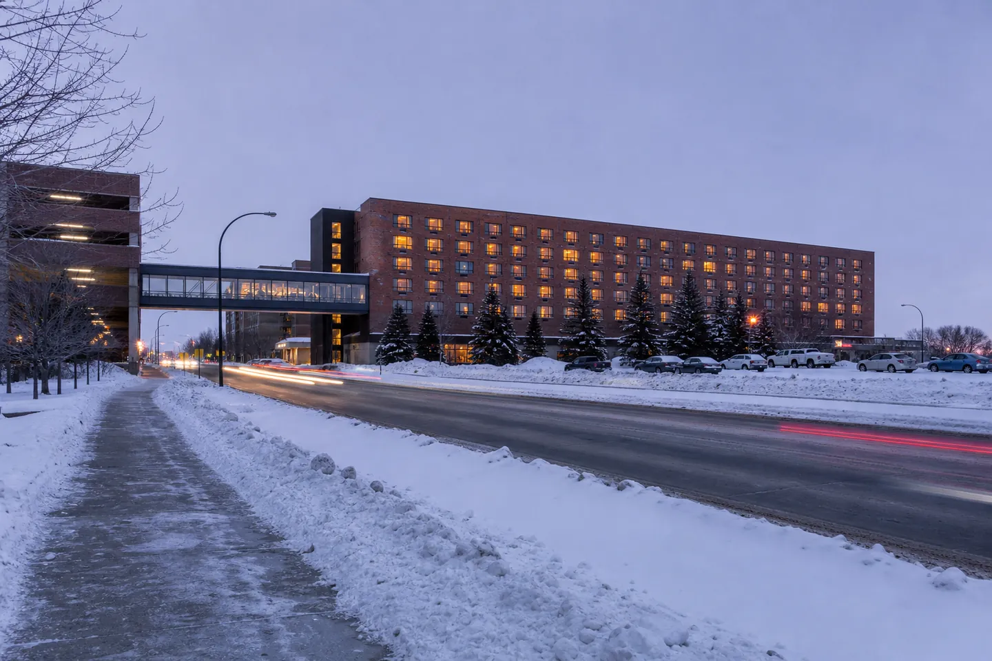 Illustration: a long brick hotel at blue hour on a snowy prairie avenue, a glass skywalk lit beside it